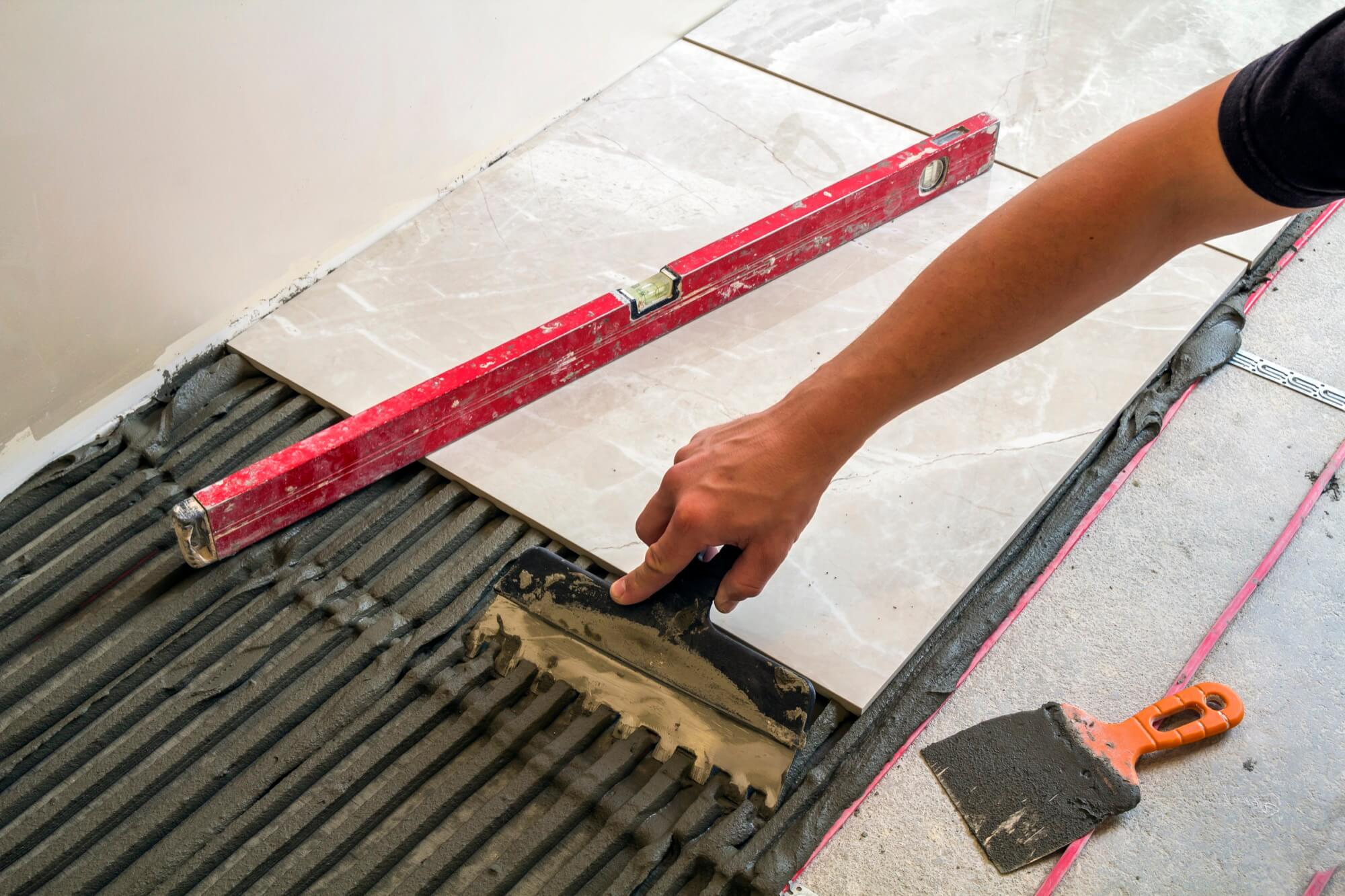 Close-up of laying tiled floor with notched trowel and spirit level during bathroom renovation.